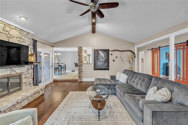 living room with lofted ceiling with beams, a stone fireplace, ceiling fan, a barn door, and dark hardwood / wood-style flooring