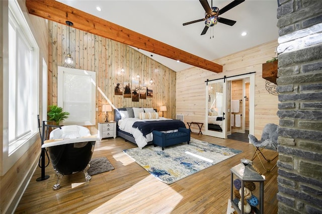 bedroom with a barn door, wooden walls, hardwood / wood-style floors, and beamed ceiling