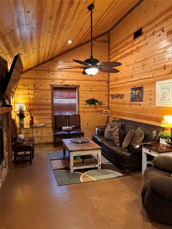 living room featuring wooden ceiling, a stone fireplace, wooden walls, ceiling fan, and concrete flooring