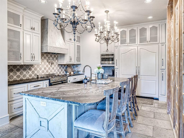 kitchen with custom range hood, sink, a notable chandelier, dark stone countertops, and an island with sink