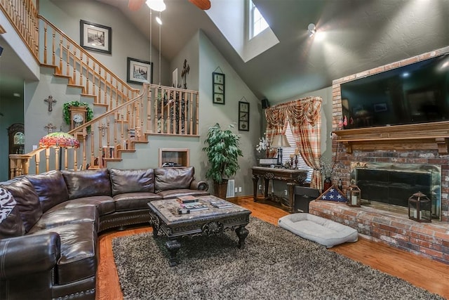 living room featuring high vaulted ceiling, a skylight, a brick fireplace, ceiling fan, and wood-type flooring