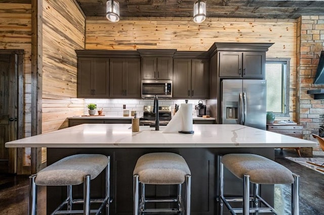 kitchen featuring a kitchen bar, appliances with stainless steel finishes, dark brown cabinets, wooden walls, and a large island with sink