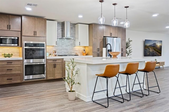 kitchen featuring wall chimney exhaust hood, light hardwood / wood-style flooring, an island with sink, a breakfast bar, and appliances with stainless steel finishes