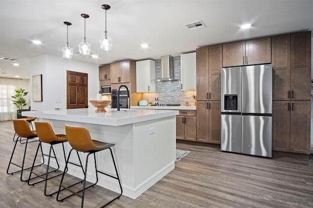 kitchen featuring pendant lighting, a kitchen island with sink, wall chimney range hood, light wood-type flooring, and appliances with stainless steel finishes