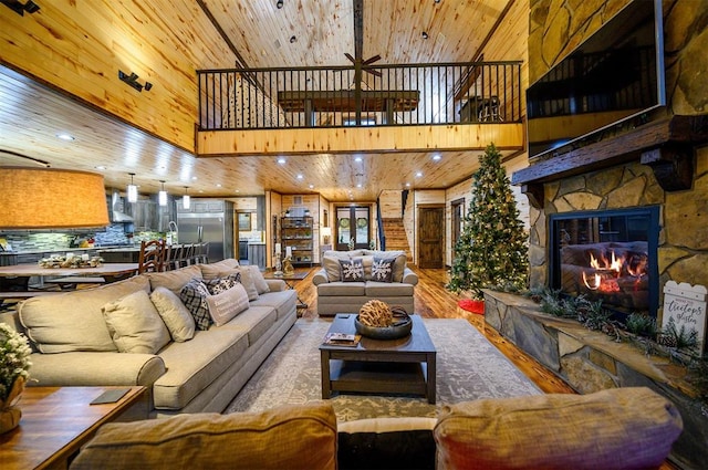 living room with a towering ceiling, ceiling fan, wood-type flooring, wooden ceiling, and a stone fireplace