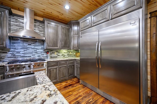 kitchen with high end appliances, wall chimney range hood, dark wood-type flooring, and tasteful backsplash