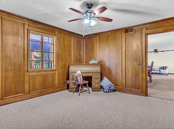 carpeted home office featuring wooden walls, ceiling fan, and a textured ceiling