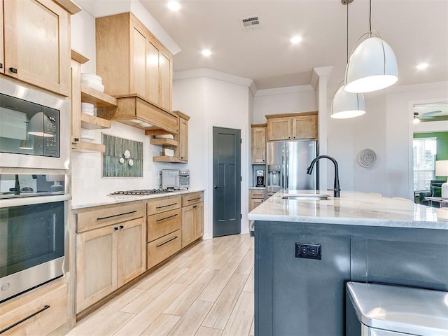 kitchen featuring appliances with stainless steel finishes, light brown cabinetry, sink, hanging light fixtures, and a center island with sink