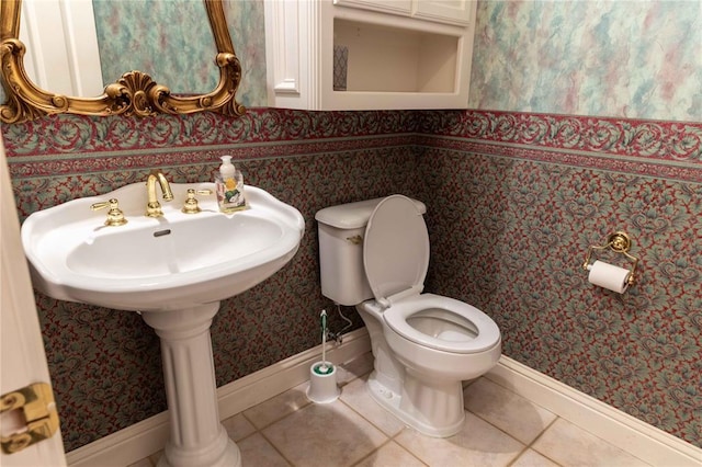 half bath featuring toilet, a sink, baseboards, tile patterned floors, and wallpapered walls