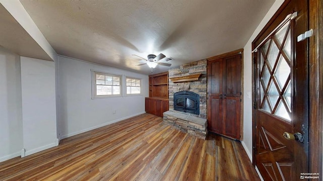 unfurnished living room with wood-type flooring, built in shelves, a stone fireplace, a textured ceiling, and ceiling fan