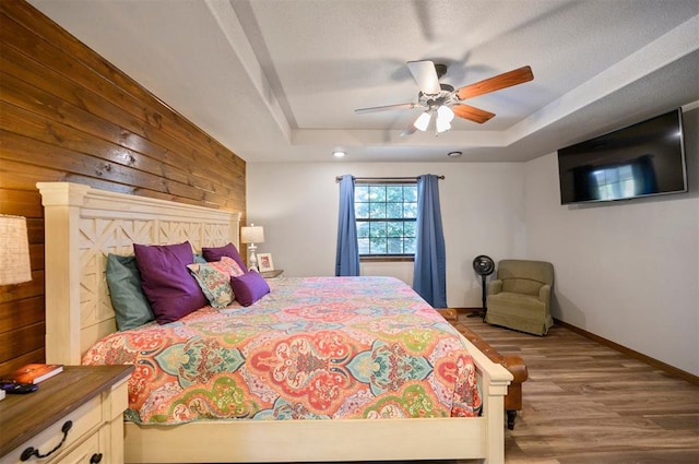 bedroom featuring a raised ceiling, light hardwood / wood-style flooring, ceiling fan, and wooden walls