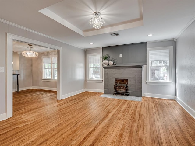 unfurnished living room with a wood stove, an inviting chandelier, a raised ceiling, light wood-type flooring, and ornamental molding