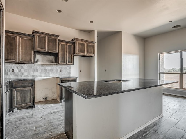 kitchen with tasteful backsplash, dark stone counters, wood finish floors, a kitchen island with sink, and dark brown cabinets