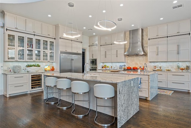 kitchen with beverage cooler, wall chimney range hood, built in appliances, white cabinets, and an island with sink