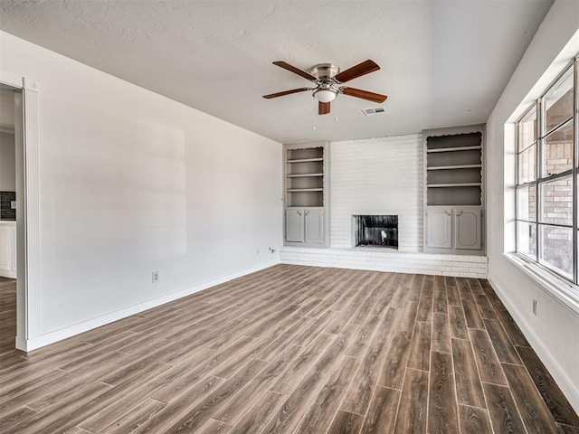 unfurnished living room featuring ceiling fan, built in shelves, dark wood-type flooring, and a brick fireplace