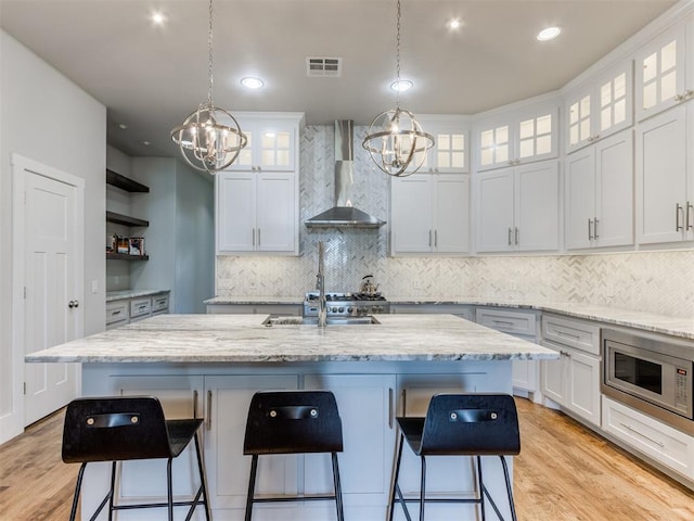 kitchen featuring white cabinets, stainless steel microwave, wall chimney range hood, and an island with sink