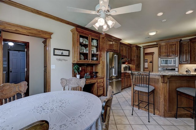 kitchen with appliances with stainless steel finishes, a kitchen breakfast bar, light stone counters, ornamental molding, and light tile patterned floors