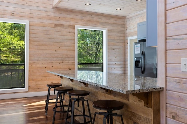 bar with wood walls, dark wood-type flooring, stainless steel refrigerator with ice dispenser, light stone counters, and wood ceiling