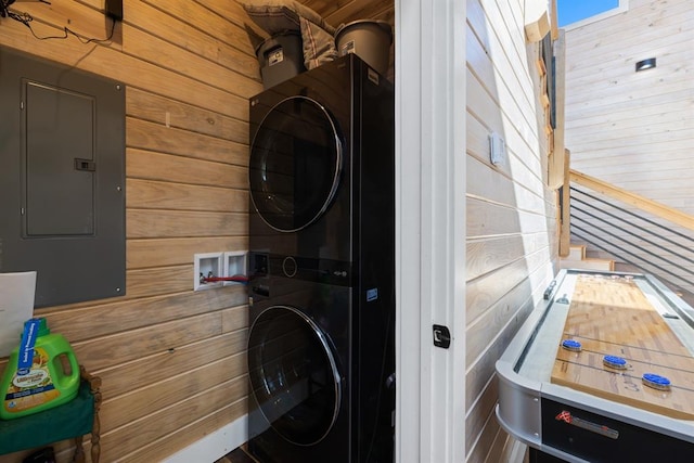 laundry area featuring electric panel, stacked washer / dryer, and wood walls