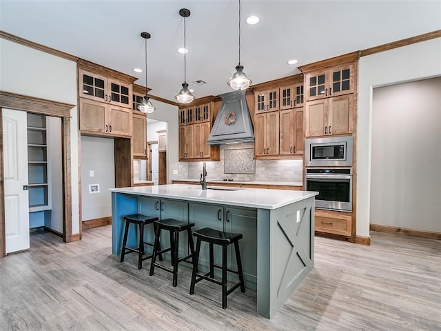 kitchen featuring custom exhaust hood, stainless steel appliances, a kitchen island with sink, pendant lighting, and light hardwood / wood-style floors