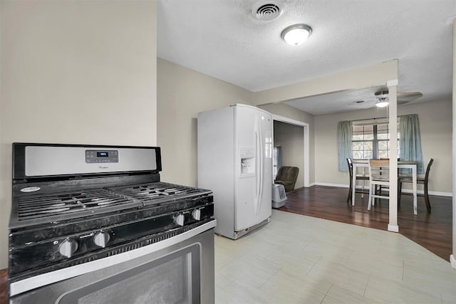 kitchen with light wood-type flooring, a textured ceiling, ceiling fan, white refrigerator with ice dispenser, and stainless steel gas stove