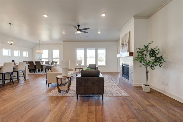 living room featuring ornamental molding, a brick fireplace, wood finished floors, baseboards, and ceiling fan with notable chandelier