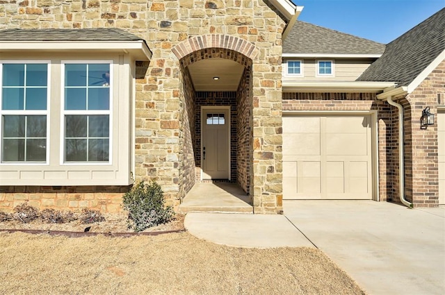 entrance to property with stone siding, roof with shingles, concrete driveway, brick siding, and a garage