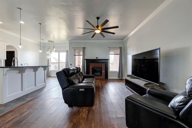 living room featuring crown molding, dark hardwood / wood-style flooring, ceiling fan with notable chandelier, and a brick fireplace