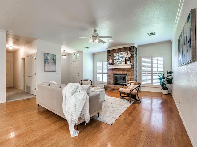 living room with a brick fireplace, a textured ceiling, ceiling fan, crown molding, and light hardwood / wood-style flooring