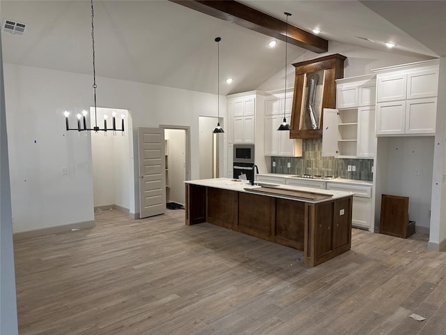 kitchen with beam ceiling, stainless steel microwave, decorative light fixtures, a kitchen island with sink, and light wood-type flooring