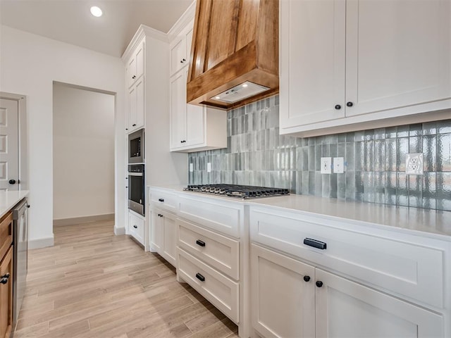 kitchen featuring light wood finished floors, custom exhaust hood, tasteful backsplash, light countertops, and appliances with stainless steel finishes