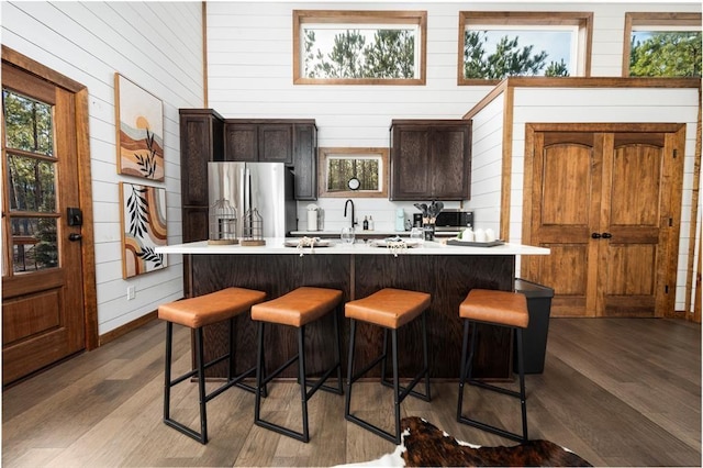 kitchen featuring stainless steel fridge, a breakfast bar, dark brown cabinetry, wood-type flooring, and wood walls