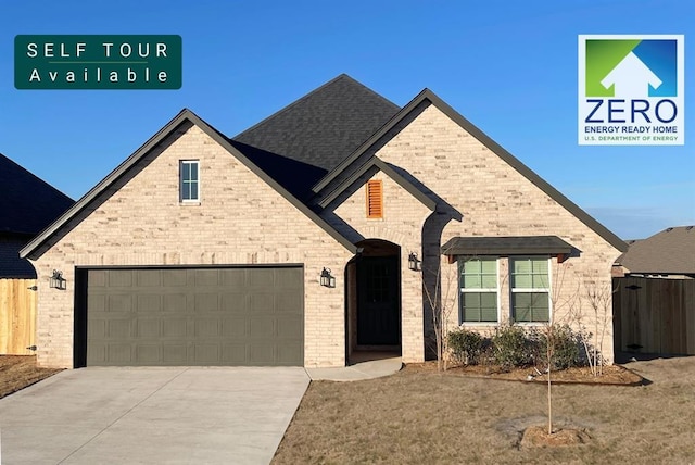 french country home with a shingled roof, concrete driveway, brick siding, and fence