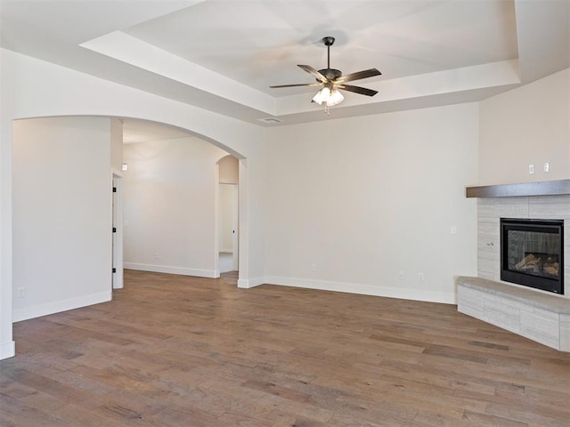 unfurnished living room with hardwood / wood-style flooring, ceiling fan, a tray ceiling, and a tiled fireplace