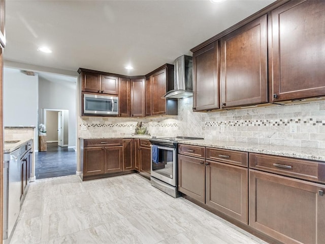 kitchen featuring light stone counters, dark brown cabinetry, appliances with stainless steel finishes, wall chimney range hood, and decorative backsplash