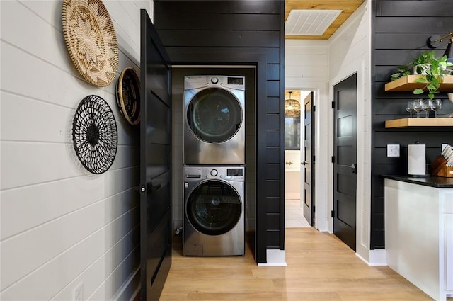 laundry room with light wood-type flooring, stacked washing maching and dryer, and wooden walls