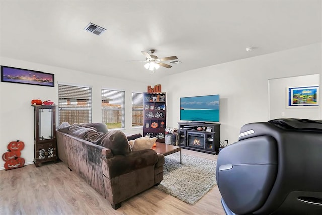 living room featuring light wood-style flooring, visible vents, ceiling fan, and a glass covered fireplace