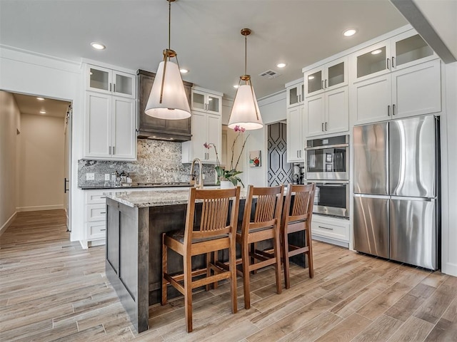 kitchen with a center island with sink, hanging light fixtures, light hardwood / wood-style flooring, light stone countertops, and stainless steel appliances