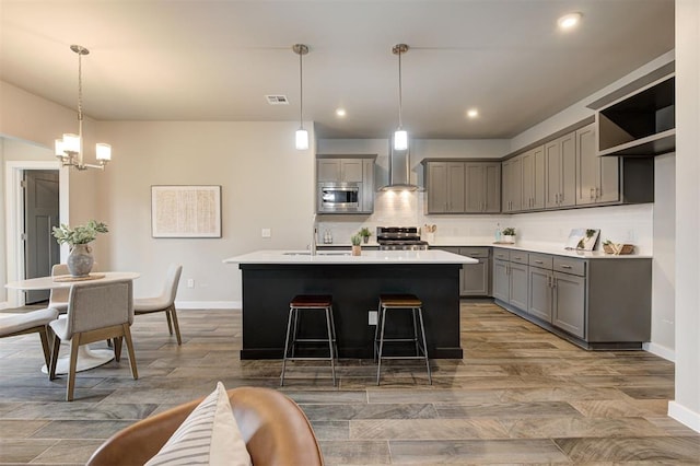 kitchen with decorative light fixtures, wall chimney range hood, stainless steel appliances, an island with sink, and gray cabinetry