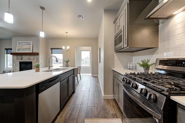 kitchen featuring wall chimney exhaust hood, stainless steel appliances, an island with sink, sink, and hanging light fixtures