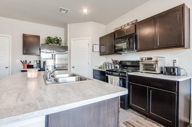 kitchen featuring appliances with stainless steel finishes, dark brown cabinetry, a kitchen island with sink, sink, and light hardwood / wood-style flooring