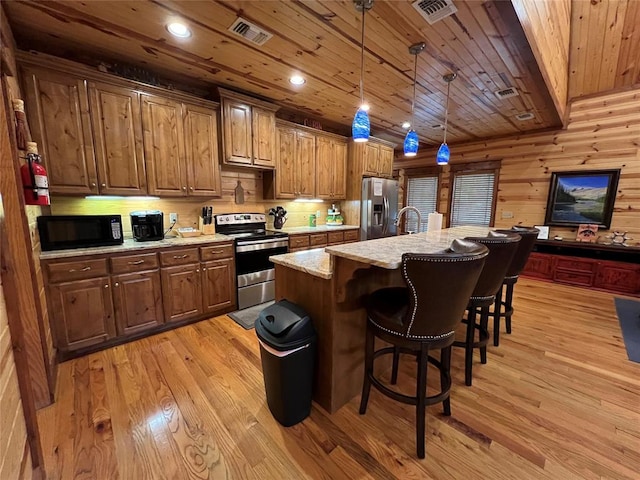 kitchen with hanging light fixtures, stainless steel appliances, wooden walls, a center island with sink, and light wood-type flooring