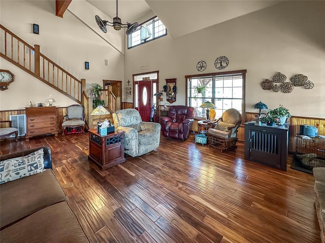 living area featuring dark wood-style flooring, beam ceiling, stairway, a high ceiling, and wainscoting