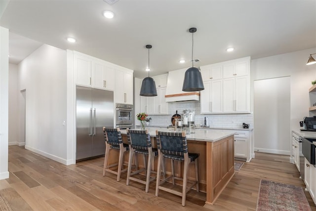 kitchen featuring built in fridge, light hardwood / wood-style flooring, pendant lighting, a kitchen island with sink, and white cabinets