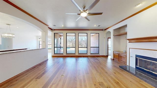 unfurnished living room featuring a tile fireplace, ceiling fan, built in shelves, ornamental molding, and light hardwood / wood-style floors