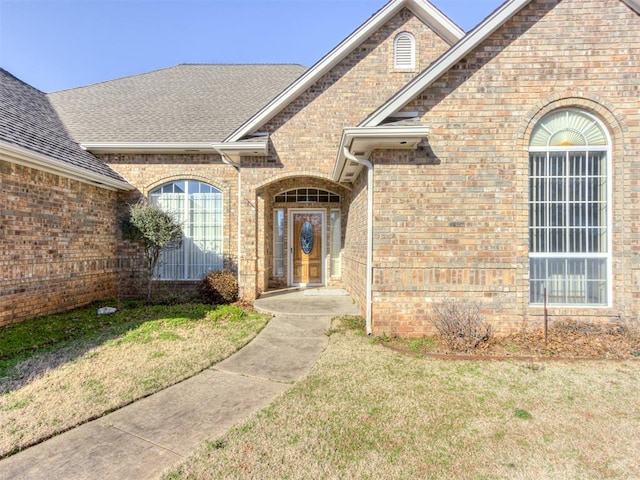 view of exterior entry featuring a yard, brick siding, and a shingled roof