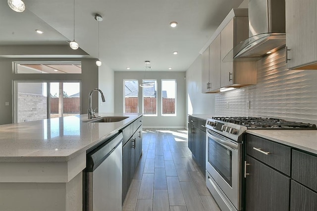 kitchen featuring appliances with stainless steel finishes, wall chimney exhaust hood, a kitchen island with sink, sink, and decorative light fixtures