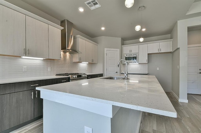 kitchen featuring appliances with stainless steel finishes, a kitchen island with sink, sink, wall chimney range hood, and pendant lighting