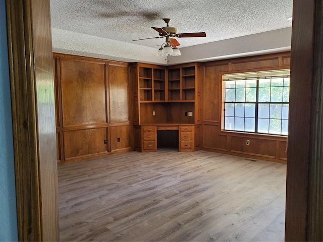 unfurnished office with wooden walls, light hardwood / wood-style flooring, ceiling fan, and built in desk