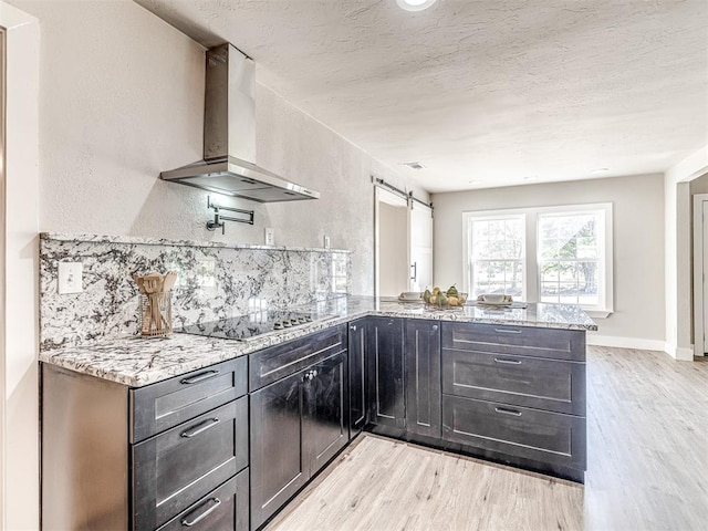 kitchen with a barn door, black electric cooktop, light stone counters, and wall chimney exhaust hood